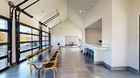 Located at 11 Wisdom Way in Seneca, SC, this single-floor event and dining space offers a contemporary open-concept design that is both functional and visually striking. The room is defined by dramatic vaulted ceilings that rise to impressive heights, amplifying the sense of openness throughout the space. Large-format gray tile flooring runs the length of the interior, providing a clean, modern foundation.

A standout architectural feature is the set of full-height glass garage doors along one wall, which flood the space with abundant natural light and offer clear views of the surrounding outdoor area and neighboring streetscape. These doors also allow the space to open up seamlessly to the exterior when desired.

The dining area is anchored by a long communal table with cross-back chairs, accommodating sizeable gatherings. Adjacent to the dining zone is a white tile-topped bar counter with industrial-style black stools, ideal for casual seating or serving. A recessed nook along the back wall provides additional counter space, suitable for buffet-style setups or equipment placement.

A smaller seating vignette near the glass doors features accent chairs alongside tropical greenery, adding warmth to the otherwise minimal aesthetic. Recessed ceiling lighting supplements the natural light, ensuring the space remains bright and welcoming throughout the day. This well-appointed venue is well-suited for group dining, events, and community gatherings.
