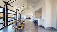 Located at 11 Wisdom Way in Seneca, SC, this single-floor event and dining space offers a contemporary open-concept design that is both functional and visually striking. The room is defined by dramatic vaulted ceilings that rise to impressive heights, amplifying the sense of openness throughout the space. Large-format gray tile flooring runs the length of the interior, providing a clean, modern foundation.

A standout architectural feature is the set of full-height glass garage doors along one wall, which flood the space with abundant natural light and offer clear views of the surrounding outdoor area and neighboring streetscape. These doors also allow the space to open up seamlessly to the exterior when desired.

The dining area is anchored by a long communal table with cross-back chairs, accommodating sizeable gatherings. Adjacent to the dining zone is a white tile-topped bar counter with industrial-style black stools, ideal for casual seating or serving. A recessed nook along the back wall provides additional counter space, suitable for buffet-style setups or equipment placement.

A smaller seating vignette near the glass doors features accent chairs alongside tropical greenery, adding warmth to the otherwise minimal aesthetic. Recessed ceiling lighting supplements the natural light, ensuring the space remains bright and welcoming throughout the day. This well-appointed venue is well-suited for group dining, events, and community gatherings.