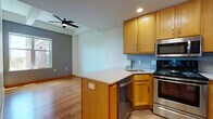 This single-floor unit offers a thoughtfully designed open-concept layout that seamlessly connects the kitchen and living areas. The kitchen is a functional centerpiece, featuring warm honey-toned oak cabinetry, a light blue subway tile backsplash, granite-style countertops, and a full suite of stainless steel appliances including a refrigerator, electric range, over-the-range microwave, and dishwasher. A peninsula island provides additional prep space and casual seating, with recessed and flush-mount lighting illuminating the area. The adjoining living space is finished with rich hardwood flooring that extends throughout the main areas, complemented by a ceiling fan, a bold slate-gray accent wall, and a window that brings in natural light and views of the surrounding building exterior. A hallway connects to additional rooms, including a carpeted space that offers flexibility as a bedroom or den. The bathroom is finished with dark espresso cabinetry, a granite-top vanity, a framed mirror, and tile flooring, presenting a clean and modern aesthetic. Overall, the space delivers a cohesive, warm, and practical interior suited for comfortable everyday living.