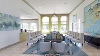 This single-story lounge space makes a strong first impression with its soaring ceilings and bright, contemporary design. Tall arched windows trimmed in bold chartreuse green line the main wall, flooding the interior with natural light while offering views of palm trees and lush outdoor surroundings, creating a seamless connection between interior and exterior. Recessed lighting and a decorative chandelier pendant complement the abundant daylight throughout the space.

The layout is thoughtfully arranged for both social interaction and relaxed seating. A central grouping features a dark upholstered sofa adorned with an array of decorative pillows in teal, navy, and green, flanked by sleek, modern swivel chairs in light gray. Along both sides of the room, rows of high-back upholstered chairs with cushioned armrests provide additional structured seating, giving the space a lounge or hospitality-oriented feel. A patterned area rug in shades of blue anchors the central seating arrangement.

The overall ambiance is polished and welcoming, blending neutral tones with vibrant accent colors to create an energetic yet comfortable atmosphere suited for gathering and relaxation.