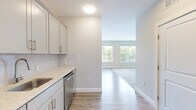 Located at 5 Lakeshore Center in Bridgewater, MA, this one-bedroom, one-bathroom studio unit delivers a clean, modern aesthetic throughout its thoughtfully designed single-floor layout. Light-toned luxury vinyl plank flooring flows through the main living and kitchen areas, complemented by soft carpet in the spacious bedroom, which features large dual windows offering lush, tree-lined outdoor views that fill the room with natural light.

The kitchen is well-equipped with crisp gray shaker-style cabinetry, gleaming quartz countertops, a subway tile backsplash, and a full suite of stainless steel appliances including a refrigerator, range, microwave, and dishwasher. Neutral walls and recessed lighting lend the kitchen a bright, functional atmosphere.

The generously sized bedroom provides ample room for living and sleeping arrangements and connects to a walk-in closet with wire shelving for organized storage. The hallway leads seamlessly between all rooms, maintaining an efficient flow throughout the unit.

The bathroom features large-format tile flooring, a quartz-topped vanity with gray cabinetry, a framed mirror, wall-mounted storage cabinet, and a tub/shower combination with wood-look tile surround and brushed gold grab bars, combining accessibility with refined style.