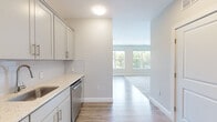 Located at 5 Lakeshore Center in Bridgewater, MA, this one-bedroom, one-bathroom studio unit delivers a clean, modern aesthetic throughout its thoughtfully designed single-floor layout. Light-toned luxury vinyl plank flooring flows through the main living and kitchen areas, complemented by soft carpet in the spacious bedroom, which features large dual windows offering lush, tree-lined outdoor views that fill the room with natural light.

The kitchen is well-equipped with crisp gray shaker-style cabinetry, gleaming quartz countertops, a subway tile backsplash, and a full suite of stainless steel appliances including a refrigerator, range, microwave, and dishwasher. Neutral walls and recessed lighting lend the kitchen a bright, functional atmosphere.

The generously sized bedroom provides ample room for living and sleeping arrangements and connects to a walk-in closet with wire shelving for organized storage. The hallway leads seamlessly between all rooms, maintaining an efficient flow throughout the unit.

The bathroom features large-format tile flooring, a quartz-topped vanity with gray cabinetry, a framed mirror, wall-mounted storage cabinet, and a tub/shower combination with wood-look tile surround and brushed gold grab bars, combining accessibility with refined style.