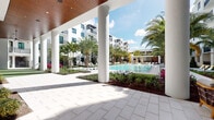 This expansive outdoor amenity area is defined by its resort-inspired design and tropical ambiance. At its center sits a generously sized swimming pool surrounded by lounge chairs and manicured landscaping, including tall palm trees, colorful tropical plants, and carefully maintained garden beds with rich mulch borders. The pool deck is finished in light-toned pavers that extend throughout the space, lending a clean, cohesive aesthetic.

A covered colonnade runs along one side of the courtyard, featuring striking white cylindrical columns and a warm wood-paneled ceiling with recessed lighting. Beneath this structure, multiple seating arrangements offer comfortable outdoor lounging, with woven armchairs and accent tables positioned to overlook the pool and greenery. A ceiling fan provides additional comfort in the shaded areas.

A poolside bar area features a sleek white countertop with woven bar stools and large fold-open windows that frame direct views of the pool and surrounding landscape. An adjacent open-air courtyard nook provides additional seating with wooden high-top tables and stools nestled among low tropical plantings. The entire space is framed by a modern multi-story building with balconies, creating a cohesive and polished outdoor retreat bathed in abundant natural sunlight.