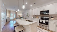 This single-floor residence showcases a cohesive coastal aesthetic with light wood flooring, neutral tones, and carefully considered finishes throughout. The kitchen serves as a functional centerpiece, featuring white shaker cabinetry, granite countertops with subtle veining, a taupe tile backsplash, and a full suite of stainless steel appliances including a range, microwave, and refrigerator. A spacious island with an undermount sink and pendant lighting above provides additional prep space and casual seating. The kitchen flows seamlessly into an open living area, where natural light enters through glass French doors framed by floor-length curtains, offering outdoor views of lush tropical greenery.

The bedrooms are carpeted and generously sized, each benefiting from windows with outdoor palm views and ceiling fans. A notable walk-in closet features wire shelving systems and ample hanging space. The bathroom includes a tub-shower combination, a vanity with granite countertops, grey cabinetry, and wood-look flooring. Throughout the residence, the design maintains a consistent coastal palette of whites, soft blues, and warm natural textures, creating a calm and inviting atmosphere.