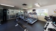 This fitness center is a dedicated, single-floor exercise facility designed to support a well-rounded workout routine. The space features durable rubber flooring throughout, providing a safe and functional surface for all types of training activities.

The cardio section is equipped with multiple treadmills and elliptical machines, including Precor-brand equipment, allowing for a variety of aerobic workouts. A wall-mounted flat-screen television is positioned for easy viewing during cardio sessions. Natural light filters in through several windows, creating a bright and energizing atmosphere.

For strength training, the facility includes a cable crossover machine, a set of free weights and dumbbells neatly organized on a rack, a weight bench, and additional resistance equipment. A large wall mirror spans a portion of the space, enhancing the sense of openness and allowing users to monitor their form during workouts.

A dedicated stretching and functional fitness area features a rack of medicine balls and stability balls in various sizes. Motivational signage is displayed on the walls, contributing to an encouraging workout environment.

Large glass doors open directly to an outdoor pool and patio area, seamlessly connecting the indoor fitness space with outdoor amenities. The center is finished with a textured ceiling and recessed fluorescent lighting, maintaining a clean and practical interior aesthetic.