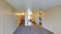 This single-floor unit presents an open and functional layout designed to maximize usability across its main living areas. The front entry is finished with wood-style flooring and features a classic six-panel door in a rich reddish-brown tone, providing a welcoming first impression. A baseboard heating unit runs along the entryway wall, offering practical climate control.

The living area extends from the entry and is covered in neutral-toned carpet, creating a comfortable and cohesive feel throughout. Structural columns define the space subtly without closing it off, maintaining an open sightline from the front door through to the rear of the unit. The walls are finished in a warm beige tone, complemented by a textured popcorn ceiling that spans the entire unit.

Toward the rear, the kitchen is partially visible and features dark lower cabinetry alongside wood-style flooring consistent with the entryway. A dedicated laundry area is accessible from the main space and houses a full-size washer and dryer, adding notable convenience. The layout balances living, cooking, and utility functions efficiently within a compact and well-organized single-level footprint.