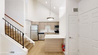 Located at 100 Maple Rd in Buffalo, NY, this two-floor residence offers a thoughtfully designed layout spread across two levels. The main floor features an open-concept kitchen and living area highlighted by dramatically vaulted ceilings that amplify the sense of space and airiness. The modern kitchen is equipped with stainless steel appliances, including a refrigerator, dishwasher, microwave, and range, complemented by light wood cabinetry, a white quartz peninsula countertop, and a pendant light fixture overhead. Light wood-tone flooring flows throughout, adding warmth to the bright, white interior. The living room centers around a tile-surround fireplace with a classic white mantel, a ceiling fan with integrated lighting, and a built-in wall niche — an inviting space bathed in natural light from a front-facing window. A French-door entry leads to a connected area with direct access to one of the two full bathrooms, each featuring tiled tub-shower combinations and wood-toned vanities with white countertops. A staircase with wood handrail and black iron balusters leads to the upper level, where a cozy room with sloped ceilings and a large window offers pleasant views of the surrounding greenery. A closet completes the home's functional layout.