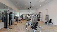 Located at 21595 Box Springs Rd in Moreno Valley, California, this single-floor fitness center is thoughtfully designed to support a comprehensive range of workout routines. The facility is organized into distinct training zones, each tailored to a specific discipline, and connected through sleek black-framed glass partition walls that maintain an open, airy feel while providing visual separation between spaces.

The strength training area features vaulted ceilings with recessed lighting and pendant ceiling fans, creating a bright and comfortable environment. Precor cable machines, weight benches, a functional trainer, and a full dumbbell rack line the room, complemented by floor-to-ceiling mirrors that amplify the sense of space. Patterned, neutral-toned carpet tiles cover the floors throughout, adding a modern, textural aesthetic.

The cardio zone is lined with large black-framed windows overlooking the exterior, flooding the space with natural light. A row of treadmills faces the windows, offering an engaging view during workouts. Adjacent to this area, elliptical trainers and stationary bikes provide additional low-impact cardio options.

A dedicated functional fitness area houses battle ropes, kettlebells, medicine balls, and a heavy punching bag with speed bag attachment, all neatly organized on wall-mounted storage racks. A separate glass-enclosed studio, visible through the partitions, accommodates cycling equipment and group fitness activities, bathed in natural light from arched windows.