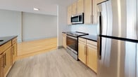 This single-floor unit showcases a thoughtfully designed open-concept layout that seamlessly connects the living and kitchen areas, creating a bright and expansive feel throughout. Warm-toned hardwood flooring runs across the main living space, complementing the clean white walls and recessed lighting overhead. Pendant lights add visual interest above the central kitchen island, which features a dark granite countertop and an integrated sink with a dishwasher. The kitchen is further equipped with light maple cabinetry, a classic white subway tile backsplash, dark granite countertops, and a full suite of stainless steel appliances including a refrigerator, range, and over-the-range microwave. Light-toned tile flooring distinguishes the kitchen zone from the hardwood living area. Large black-framed windows and sliding glass doors at the far end of the living space invite abundant natural light and provide access to a balcony, offering open outdoor views. The bathroom features a neutral tile floor, partial tile wainscoting, a white vanity with dark countertop, a large mirror, and a separate enclosed shower area, maintaining the unit's consistent modern aesthetic.