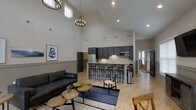 Located at 5020 Garrett Rd in Durham, NC, this single-floor space makes a strong architectural impression with its dramatic vaulted ceilings that soar overhead, amplifying the sense of openness throughout. Natural light filters in through high clerestory windows, complementing the recessed lighting and a statement chandelier that anchors the main gathering area. The open-concept layout seamlessly connects the lounge and kitchen zones, creating a fluid and functional environment. The lounge area is appointed with a sleek leather sofa, rustic wood accent tables, and a large area rug, all set against warm-toned hardwood flooring. A framed piece of abstract artwork adds a curated, modern touch to the neutral wall palette. The kitchen is well-equipped with dark cabinetry, stainless steel appliances including a full-size refrigerator and microwave, and a generous island counter with granite-style surfaces and bar-height seating for multiple guests. This bar area serves as a natural social hub within the space. A hallway visible in the background suggests additional rooms or access points, and a glass-paneled door allows views to an exterior area, further enhancing the bright, airy atmosphere throughout.