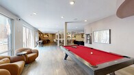 Located at 1600 W Call St in Tallahassee, Florida, this thoughtfully designed single-floor amenity space offers a diverse range of social and recreational zones within a generously sized footprint. The open-concept main area features wide-plank wood-look flooring, exposed ceiling beams, and floor-to-ceiling windows along one side that invite abundant natural light and views of the surrounding exterior. A full-size pool table with a bold red felt surface anchors the game zone, complemented by an air hockey table nearby. Adjacent lounge seating — including curved caramel-toned accent chairs and a large sectional leather sofa paired with a marble-top coffee table — provides comfortable gathering spots throughout. Textured accent walls with subtle linear patterns add visual depth and a modern character to the space. A well-appointed kitchenette serves the area with warm-toned cabinetry, a beverage cooler, microwave, sink, and open shelving. A dedicated media lounge features a large-screen display, moody ceiling lighting, patterned carpet, and curated artwork with a theater theme, creating an immersive retreat. A more intimate sitting area with leather accent chairs and sophisticated décor rounds out this multifunctional community hub, making it an ideal space for socializing, recreation, and relaxation.
