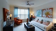 This single-floor space offers a comfortable and thoughtfully designed living environment. The living room serves as the centerpiece, showcasing a welcoming atmosphere with a carefully curated color scheme that blends warm orange and rust tones with cool aqua and blue accents. A large window dressed with light blue grommet curtains and horizontal blinds allows generous natural light to fill the room, creating a bright and airy feel throughout the day. A ceiling fan with integrated lighting is centrally mounted overhead, providing both comfort and practical illumination. The space is furnished with a plush white sofa adorned with decorative pillows in coordinating patterns, complemented by a rich cognac leather accent chair that adds a bold, stylish contrast. A sleek dark wood coffee table anchors the seating arrangement, while a coordinating console table along the wall provides additional surface space for decorative accents. A vibrant large-scale floral canvas artwork adds a striking visual focal point to the wall. Soft wall-to-wall carpeting underfoot enhances the overall sense of warmth and coziness, making this a practical and visually appealing living space.