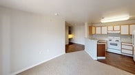 This single-floor residential unit offers a practical and well-organized layout designed for comfortable everyday living. The open-concept living area features neutral-toned carpet, clean white walls, and a sliding glass door that invites natural light and provides outdoor views. The layout connects seamlessly to the kitchen, which is equipped with a full suite of white appliances including an electric range, refrigerator, and dishwasher. Warm wood-toned cabinetry lines the kitchen walls, complemented by light speckled countertops and a double-basin sink, offering ample storage and counter space for meal preparation. A breakfast bar-style counter further connects the kitchen to the living space, enhancing the open feel of the common areas. The bedrooms are carpeted and finished with neutral grey walls, each featuring vertical blind-covered windows that allow for adjustable natural light. The bathrooms are appointed with wood-style vinyl flooring, providing a clean contrast to the lighter tones found in the rest of the unit. The primary bathroom includes a vanity with wood cabinetry, a large mirror with overhead lighting, and a full tub-shower combination. A second bathroom offers a similarly functional design with a tub-shower unit and a toilet. Throughout the unit, consistent neutral finishes and thoughtful connectivity between rooms create a cohesive and livable environment.