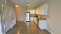 This single-floor space showcases an open-concept design that seamlessly integrates the kitchen and living areas, creating a sense of spaciousness and functionality. The kitchen is a standout feature, outfitted with stainless steel appliances, including a full-size refrigerator, electric range, built-in microwave, and dishwasher. Crisp white cabinetry lines the walls, providing ample storage, while granite countertops add a touch of elegance. A undermount sink with a modern faucet completes the kitchen workspace. The flooring throughout is a rich, dark hardwood, lending warmth and continuity to the open layout. Neutral-toned walls complement the clean, contemporary aesthetic, while track lighting above the kitchen and a flush-mount ceiling fixture ensure well-distributed illumination. The entryway, visible from the main living space, features a paneled door with a louvered accent, adding a classic architectural detail. The layout's open flow between the kitchen and living area makes the space feel expansive and well-suited for both everyday living and entertaining.