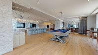 This generously scaled single-floor amenity space is designed for upscale entertaining, seamlessly blending a variety of functional areas within an open-concept layout. Wide-plank hardwood flooring flows throughout, unifying each zone and enhancing the airy, modern aesthetic.

At the heart of the space, a full catering kitchen commands attention with an elongated island topped in white stone, stainless steel appliances, pendant lighting, and a striking geometric hexagonal tile accent wall in aqua, grey, and white tones. Dual flat-screen televisions are mounted above the countertop wall, making this area ideal for gatherings. A long bar with light blue upholstered seating extends the kitchen's entertaining capacity.

Adjacent to the kitchen, a billiard table anchors an open game area, illuminated by a cascading crystal pendant fixture. The space transitions naturally into multiple lounge areas, where curved and sectional seating arrangements are appointed with layered rugs and contemporary lighting. 

Floor-to-ceiling windows frame breathtaking panoramic ocean views, flooding the lounge areas with natural light and creating a seamless connection to the outdoors. Sheer drapery softens the expansive glazing while maintaining the coastal ambiance. Textured stone columns and refined architectural details throughout add depth and character, completing this sophisticated, resort-style entertainment destination.