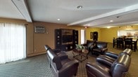 This single-floor space is designed for comfortable communal living and gathering, seamlessly blending a lounge area and a formal dining space in an open-concept layout. The living area is anchored by a set of plush dark leather armchairs arranged around a round coffee table, creating an inviting seating arrangement ideal for conversation or relaxation. A large dark wood entertainment center with open shelving, display compartments, and a flat-screen television serves as the focal point of the lounge. Warm earth-toned walls in shades of gold and tan lend a cozy, welcoming ambiance throughout. The dining area features a substantial dark wood table surrounded by matching cross-back chairs, illuminated by an elegant multi-arm chandelier that adds a refined touch. A coordinating dark wood sideboard with ample cabinet storage lines one wall, complemented by framed artwork in rich, warm hues. Patterned carpeting flows throughout the space, tying the two areas together cohesively. Recessed lighting and a wall-mounted HVAC unit provide practical comfort, while baseboard heating ensures year-round warmth. The overall design balances functionality with a welcoming, furnished aesthetic suited for everyday use and social gatherings.