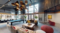 Located at 2200 W Meeker St in Kent, Washington, this single-floor community clubhouse showcases a modern industrial design characterized by soaring ceilings, expansive floor-to-ceiling windows, and a bold palette of dark steel, warm wood accents, and reclaimed wood wall panels. The entry foyer welcomes visitors through sleek glass doors framed in matte black, accented by potted greenery and framed artwork, establishing a polished first impression.

The main lounge area is generously proportioned, anchored by an arrangement of plaid armchairs, a tufted dark leather sofa, and textural ottomans gathered around a wood coffee table. A dramatic cluster of sculptural pendant lights with layered, organic forms hangs overhead, adding artistic character to the space. A large flat-screen display and comfortable seating create a dedicated media zone, while reclaimed wood panels lend warmth against the dark upper walls.

The game area features a pool table and foosball table positioned alongside oversized windows that flood the space with natural light and offer views of the surrounding exterior. Adjacent to this is a well-appointed kitchen bar with light wood flooring, a sleek island with white barstools, open shelving, pendant lighting, and a dining area—ideal for casual gatherings. Throughout, the open layout and abundant glazing create a bright, energetic, and welcoming communal atmosphere.
