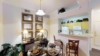 This single-floor residence offers a thoughtfully designed layout that balances style and functionality. The dining area serves as a focal point, featuring a round glass-top dining table surrounded by upholstered chairs, set beneath a classic pendant light fixture. Crown molding along the ceiling adds a refined architectural detail that elevates the overall aesthetic. The walls are adorned with a curated gallery of gold-framed artwork, complemented by warm table lamps and lush greenery that create an inviting, lived-in atmosphere. A pass-through opening connects the dining space to the kitchen, providing an open and airy feel that encourages interaction between the two areas. The kitchen showcases light wood cabinetry, white appliances, and a breakfast bar with bar seating — ideal for casual dining or entertaining. Upper shelving above the cabinetry displays decorative pottery and plants, adding vertical interest and charm. Soft, neutral wall tones throughout the space lend a warm and cohesive ambiance, while natural and artificial lighting work together to keep the interior bright and welcoming. The overall design aesthetic is classic and elegant, with carefully chosen furnishings and decor that give the space a polished, comfortable character.