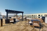 Located atop 90 Ocean Ave in Revere, MA, this rooftop deck offers a well-appointed outdoor amenity space with sweeping open-air views of the surrounding area. The deck is finished with warm wood-tile flooring laid in a parquet-style pattern, lending a refined aesthetic to the expansive terrace. A striking dark-framed pergola serves as the architectural centerpiece, providing partial shade over a curated lounge area furnished with rattan hanging chairs and wicker seating. Dining tables and chairs are positioned throughout the deck, ideal for al fresco gatherings, while grill stations are available for outdoor cooking. The terrace is enclosed by a sleek perimeter railing, ensuring safety while preserving unobstructed sightlines beneath open skies. Access to the rooftop is provided through a bright, modern hallway featuring light hardwood-style flooring, a stainless steel elevator set against a bold blue vertical-slat accent wall, and floor-to-ceiling glass walls that flood the space with natural light. A separate room off the hallway features a striking horizontal-stripe wallcovering and three tall arched mirrors, adding a stylish, functional touch. Together, the interior and exterior elements create a cohesive, elevated amenity experience.