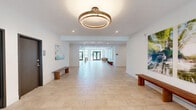 Located at 225 Cherokee St in Bethlehem, PA, this single-floor amenity space offers a thoughtfully designed environment that blends comfort, functionality, and contemporary aesthetics. The interior is finished with large-format light stone tile flooring throughout, crisp white walls, and recessed lighting, creating a clean and welcoming atmosphere.

The living lounge area serves as the social centerpiece of the space, furnished with tan leather sofas, a wood-and-metal coffee table, and a wall-mounted flat-screen TV. Nature-inspired artwork adorns the walls, adding warmth and color to the modern palette. Floor-to-ceiling black-framed windows flood the lounge with natural light and frame views of the surrounding greenery.

A glass-enclosed fitness area is seamlessly integrated into the layout, offering visual openness while providing a dedicated workout zone equipped with a dumbbell rack, weight bench, stability ball, and cardio equipment. The transparent walls maintain the sense of spaciousness across the entire floor.

The entryway greets visitors with a generous open foyer featuring large glass double doors that open directly to an outdoor patio, inviting abundant natural light inside. A comfortable accent chair and curated artwork give the entrance a polished, residential feel. Wooden benches along the open corridor add practical seating throughout the communal areas, completing this well-appointed amenity space.