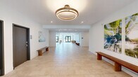 Located at 225 Cherokee St in Bethlehem, PA, this single-floor amenity space offers a thoughtfully designed environment that blends comfort, functionality, and contemporary aesthetics. The interior is finished with large-format light stone tile flooring throughout, crisp white walls, and recessed lighting, creating a clean and welcoming atmosphere.

The living lounge area serves as the social centerpiece of the space, furnished with tan leather sofas, a wood-and-metal coffee table, and a wall-mounted flat-screen TV. Nature-inspired artwork adorns the walls, adding warmth and color to the modern palette. Floor-to-ceiling black-framed windows flood the lounge with natural light and frame views of the surrounding greenery.

A glass-enclosed fitness area is seamlessly integrated into the layout, offering visual openness while providing a dedicated workout zone equipped with a dumbbell rack, weight bench, stability ball, and cardio equipment. The transparent walls maintain the sense of spaciousness across the entire floor.

The entryway greets visitors with a generous open foyer featuring large glass double doors that open directly to an outdoor patio, inviting abundant natural light inside. A comfortable accent chair and curated artwork give the entrance a polished, residential feel. Wooden benches along the open corridor add practical seating throughout the communal areas, completing this well-appointed amenity space.