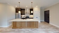 Located at 525 Ridge St in Charlottesville, Virginia, this single-floor residence combines modern design with high-quality finishes in a clean, contemporary aesthetic. Light wood-tone luxury flooring runs throughout the entire space, creating a warm and cohesive feel from room to room.

The open-concept kitchen is a true centerpiece, showcasing a large island with a natural wood base and crisp white quartz countertop, complemented by a matte black sink and globe pendant lights with brass accents. Dark shaker-style cabinetry with gold hardware lines the walls, contrasted by a stainless steel refrigerator, electric range, and a sleek wall-mounted range hood. A textured tile backsplash adds depth and visual interest.

The living area is bright and airy, with multiple windows dressed in roller shades that frame lush green tree views, and French doors that open to an outdoor deck, inviting natural light and fresh air. The bedrooms continue the light, neutral palette with soft gray walls, recessed lighting, and ceiling fans for year-round comfort.

The bathroom is finished with floor-to-ceiling white rectangular tile in the walk-in shower, accented by a hexagonal mosaic niche, a frameless glass door with matte black hardware, and a wood-toned vanity — all contributing to a sleek, spa-inspired atmosphere.