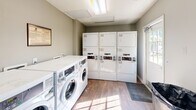 Located at 6889 Idols Rd in Clemmons, North Carolina, this shared laundry facility is a well-organized, single-room space designed for efficient, practical use. The room features commercial-grade front-loading washing machines positioned along one wall, complemented by a bank of stacked dryer units along the opposite wall, offering ample laundry capacity. The flooring throughout is a warm wood-look material that adds a clean, modern aesthetic to the utilitarian space. Neutral gray walls keep the environment feeling fresh and tidy. Natural light filters in through multiple white-framed windows fitted with horizontal blinds, brightening the interior and creating a comfortable atmosphere. A fold-down countertop along one wall provides a convenient surface for sorting or folding laundry. Safety is addressed with a mounted fire extinguisher clearly visible on the wall. Overhead fluorescent lighting ensures the space remains well-lit at all times. A security camera is also present, offering added peace of mind. A glass-paneled exterior door provides direct outdoor access, further enhancing ventilation and light. Service information is posted on the wall for easy reference. The space is compact yet thoughtfully arranged to maximize functionality.