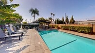 Located at 10853 Firestone Blvd in Norwalk, California, this single-level outdoor amenity space centers around a generously sized, crystal-clear swimming pool framed by a clean concrete deck. Lounge chairs are arranged along the poolside, offering ample space for sunbathing under the Southern California sun. Adjacent to the pool, a covered patio area provides welcome shade, featuring a pergola-style steel roof structure equipped with overhead speakers. Beneath it, a dining table with chairs and market umbrellas creates an ideal setting for outdoor gatherings. A built-in counter or bar surface along the edge of the covered zone adds functional utility to the space. The surrounding landscape is richly planted with banana trees, palms, hedged shrubs, and ornamental grasses, lending a tropical and private atmosphere. Decorative planters are positioned throughout the patio, enhancing the greenery. A perimeter wall lined with trimmed hedges and masonry provides privacy and enclosure. The overall ambiance is bright, open, and resort-inspired, well-suited for relaxation and social use in a warm, sun-filled Southern California setting.