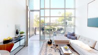 Located at 3008 Santa Monica Blvd in Santa Monica, CA, this contemporary two-story loft showcases an open, modern design with soaring double-height ceilings and polished concrete floors throughout. The main level is anchored by an expansive living area bathed in natural light from dramatic floor-to-ceiling glass windows and doors that open directly to a private outdoor patio, offering views of lush greenery and palm trees — a true California indoor-outdoor lifestyle.

The well-appointed kitchen is outfitted with sleek gray cabinetry extending to the ceiling, stainless steel appliances including a gas range, built-in microwave, full-size refrigerator, and a dark subway tile backsplash that adds a sophisticated contrast to the white quartz countertops. A dedicated dining area sits adjacent to the kitchen, ideal for everyday meals or entertaining.

The upper loft level serves as a private sleeping quarters, accessible via a staircase and overlooking the main living space below. The ground floor also includes a full bathroom with a vanity, and a discreet in-unit laundry closet equipped with a stacked full-size washer and dryer. The overall ambiance is clean, urban, and light-filled, reflecting the best of modern Southern California design.