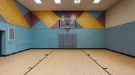 This indoor basketball court is a well-appointed recreational space designed for both competitive play and casual athletic activity. The court features a classic hardwood-style floor with clearly marked regulation lines, including a painted three-point arc and key, providing an authentic playing experience. A regulation-height glass backboard and hoop is mounted on the primary end wall, accompanied by wall-mounted protective padding beneath it for player safety.

The space is visually striking, with the main feature wall adorned in a bold geometric mural composed of large diagonal shapes in gold, burgundy, and teal — incorporating textured wood slat panels that add warmth and dimension. The lower half of the surrounding walls is painted in a cool teal-blue, creating a cohesive and energetic atmosphere throughout the court.

Soaring ceilings contribute to an open, airy feel, and are outfitted with recessed rectangular lighting panels that ensure bright, even illumination across the entire playing surface. High-mounted windows along one wall allow natural light to filter in, further enhancing the brightness of the space. An interior observation window on the side wall offers a view into an adjacent room. The court also includes multiple court markings in teal, accommodating various game formats and drills.