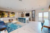 Located at 645 E Dania Beach Blvd in Dania Beach, Florida, this single-floor space delivers a sophisticated coastal-modern aesthetic throughout its well-appointed layout, encompassing a living room, kitchen, and dining room. Expansive large-format marble-style tile flooring flows seamlessly across the entire space, lending an air of elegance and continuity. The kitchen serves as a visual centerpiece, anchored by a striking blue-veined marble island with teal bar stools and pendant lighting overhead. Taupe cabinetry and a matching marble accent wall frame a wall-mounted flat-screen TV, complemented by an illuminated display cabinet. The living area features a curved navy leather sofa atop an ocean-wave area rug, with bold large-scale artwork adding a dynamic focal point. Floor-to-ceiling sliding glass doors flood the space with natural light and open directly onto an outdoor patio, offering lush greenery views. The dining area is appointed with a live-edge wood communal table surrounded by teal upholstered chairs, beneath a dramatic rustic wood beam chandelier with Edison bulb pendants. White shiplap-style walls and nautical-themed artwork complete the coastal character of this thoughtfully curated space.