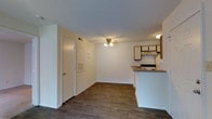 This single-floor residence offers a practical and functional layout centered around an open-concept living and kitchen area. The living space features rich, dark hardwood-style flooring that extends throughout, creating a warm and cohesive aesthetic. A ceiling fan with a light fixture is centrally positioned, providing both air circulation and ambient lighting. The walls are finished in a clean, neutral white, complemented by a textured ceiling that adds subtle visual interest.

The kitchen is seamlessly integrated into the living area via a raised breakfast bar or counter peninsula, offering a natural division between the two spaces while maintaining an open feel. The kitchen is equipped with an electric range, a range hood, a refrigerator, and wood-toned upper cabinetry with light-colored lower cabinets, providing ample storage. A wall-mounted vent and access panel are also visible near the entry, indicating well-maintained utility systems. The space is efficiently designed to maximize usability within a compact footprint, making it well-suited for comfortable everyday living.