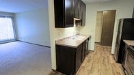 This single-story space showcases a functional kitchen that serves as a central feature of the layout. The kitchen is outfitted with dark espresso-toned cabinetry, both upper and lower, providing ample storage. A double-basin sink with a chrome faucet is set into laminate countertops that offer a neutral, earthy tone complementing the dark cabinetry. The kitchen flooring consists of wood-look laminate planks, adding warmth and durability to the space.

The kitchen opens directly to an adjacent living area, which transitions from hard flooring to carpet, creating a clear distinction between the two zones while maintaining an open, connected feel. The layout promotes easy flow between the kitchen and living space, making the area well-suited for everyday use.

A doorway visible from the kitchen leads to what appears to be a closet or additional storage area, complete with built-in shelving, adding to the overall functionality of the unit. The walls are finished in a neutral off-white tone throughout, contributing to a clean, understated aesthetic. Overall, the space is practical and straightforward in its design.