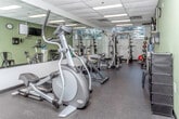 This fitness center at 4661 Woodward Ave in Detroit, Michigan, offers a well-equipped, single-floor exercise space designed to support a range of workout routines. The room features a drop ceiling with recessed panel lighting that ensures consistent, bright illumination throughout. Sage green accent walls contrast with neutral white surfaces, while large windows along one wall allow natural light to filter in and provide views of surrounding greenery.

The space is outfitted with a diverse selection of cardio equipment, including a TM6000 treadmill, an elliptical trainer, and a spin bike, catering to various cardiovascular training preferences. For strength training, the facility includes a multi-station cable machine, a power rack, an adjustable bench, and a rack of dumbbells in multiple weights. Exercise balls and medicine balls are also available for functional fitness training.

Full-length mirrors run along one wall, visually expanding the space while allowing users to monitor their form during workouts. Rubber flooring provides a durable, shock-absorbent surface suitable for both cardio and strength activities. Additional amenities include a wall-mounted flat-screen television, a water cooler, paper towel dispensers, and a clock. The overall layout is organized and functional, making efficient use of the available floor space.