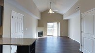This single-floor space is defined by its striking architectural design and open, airy atmosphere. The centerpiece of the main living area is a dramatic vaulted ceiling that rises to an impressive peak, enhancing the sense of spaciousness throughout. A ceiling fan is centrally mounted, providing both function and a subtle design accent against the clean, neutral-toned walls.

Rich, dark hardwood flooring flows seamlessly across the living area, adding warmth and a polished finish to the space. A corner fireplace with a sleek, modern surround serves as a focal point, offering both visual interest and a cozy gathering spot. Adjacent to the fireplace, a sliding glass door floods the room with natural light and provides direct access to an outdoor balcony, where views of the surrounding environment can be enjoyed.

From the living area, a granite-topped kitchen counter is partially visible, hinting at a well-appointed kitchen that transitions naturally into the open-concept layout. The combination of high ceilings, abundant natural light, quality finishes, and thoughtful architectural details creates an inviting and functional living environment suitable for a variety of lifestyles.