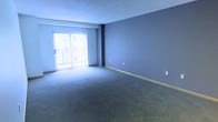 This single-floor residential unit offers a well-organized and functional layout designed for comfortable everyday living. The main living area is a generously sized, open room finished with wall-to-wall carpet in a neutral tone and painted in a cool blue-gray hue that creates a calm, airy atmosphere. A large sliding glass door fitted with vertical blinds fills the space with natural light and provides direct access to a private balcony, offering a pleasant outdoor connection. The living area is accented by clean baseboards and includes multiple electrical outlets, reflecting a practical and move-in-ready design.

The kitchen is designed in a classic galley configuration, featuring white cabinetry that lines both walls from floor to ceiling, maximizing storage capacity. Dark granite countertops provide a striking contrast against the white cabinets, lending the kitchen a sleek, modern aesthetic. Stainless steel appliances — including a refrigerator, microwave, electric range, and dishwasher — are seamlessly integrated throughout. Wood-look flooring runs the length of the kitchen, adding warmth and durability. Overhead track lighting ensures the workspace is well-lit. The thoughtful layout of this unit balances open living with efficient, functional spaces throughout.