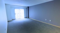 This single-floor residential unit offers a well-organized and functional layout designed for comfortable everyday living. The main living area is a generously sized, open room finished with wall-to-wall carpet in a neutral tone and painted in a cool blue-gray hue that creates a calm, airy atmosphere. A large sliding glass door fitted with vertical blinds fills the space with natural light and provides direct access to a private balcony, offering a pleasant outdoor connection. The living area is accented by clean baseboards and includes multiple electrical outlets, reflecting a practical and move-in-ready design.

The kitchen is designed in a classic galley configuration, featuring white cabinetry that lines both walls from floor to ceiling, maximizing storage capacity. Dark granite countertops provide a striking contrast against the white cabinets, lending the kitchen a sleek, modern aesthetic. Stainless steel appliances — including a refrigerator, microwave, electric range, and dishwasher — are seamlessly integrated throughout. Wood-look flooring runs the length of the kitchen, adding warmth and durability. Overhead track lighting ensures the workspace is well-lit. The thoughtful layout of this unit balances open living with efficient, functional spaces throughout.