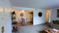 This single-floor residence offers a welcoming and functional layout designed for comfortable everyday living. The living area features neutral wall-to-wall carpeting that creates a soft, warm atmosphere, while a large decorative clock adds a distinctive accent to the otherwise clean white walls.

The space opens seamlessly into the dining area, which is distinguished by rich hardwood-style flooring that provides a natural contrast to the carpeted living room. A ceiling fan with integrated lighting fixture is centered above the dining space, providing both practical airflow and ambient illumination. A round glass-top dining table surrounded by wicker-style chairs sits at the heart of this area, giving the room a casual yet inviting character.

Just beyond the dining area, the kitchen is partially visible and features dark cabinetry alongside a full-size white refrigerator, hinting at a functional and well-equipped cooking space. Ceiling-mounted HVAC vents and floor-level registers throughout indicate a full climate control system. The open-plan design between the living, dining, and kitchen areas promotes a natural flow and makes the overall space feel airy and connected, ideal for both everyday routines and casual entertaining.