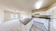 This single-floor residence offers a thoughtfully designed open-concept layout that seamlessly connects the kitchen and living areas. The kitchen is a standout feature, showcasing crisp white cabinetry, dark granite-style countertops, a central island with additional storage, and white appliances including a range, dishwasher, and refrigerator. Wood-look plank flooring runs throughout the kitchen, providing a clean, modern contrast to the warm, neutral wall tones. The living area flows naturally from the kitchen and is finished with plush carpeting, a ceiling fan with light fixture, and generous natural light from multiple windows. French doors open directly to a private balcony, offering outdoor access and views of the surrounding community. The bedrooms are carpeted and finished with the same neutral palette, providing a calm and versatile backdrop. The bathroom is well-appointed with wood-look tile flooring, a tiled tub-shower combination, a single-sink vanity with white cabinetry, and a large mirror. Clean white trim and consistent finishes throughout the space contribute to a cohesive and functional living environment that is both practical and inviting.