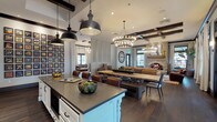 This single-floor space showcases a masterfully designed open-concept layout that seamlessly integrates a gourmet kitchen, dining area, and living lounge into one cohesive environment. Exposed dark wood ceiling beams run throughout, grounding the design with a rustic character that contrasts elegantly against crisp white walls and cabinetry.

The kitchen is a standout feature, outfitted with white shaker-style cabinetry, dark hardware, and grey stone countertops extending across a generously sized central island with turned legs. A striking antique mirror backsplash with black-framed panes reflects light and adds depth to the space. Industrial-style pendant lights hang overhead, complementing the wine cooler and integrated appliances built into the cabinetry wall. Dark hardwood flooring flows continuously throughout the entire space.

The dining area accommodates a large farmhouse-style table surrounded by leather and wood chairs, anchored beneath a multi-tiered candle-style chandelier. Adjacent, the lounge area features a full stacked-stone fireplace, leather club chairs, a wooden coffee table, and layered area rugs atop the dark hardwood floors. A large round wall clock serves as a dramatic focal point. Tall black-framed windows flood the lounge with natural light, completing the warm, sophisticated ambiance.