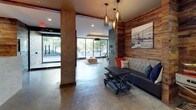This single-floor space showcases a striking industrial-modern aesthetic, blending raw materials with thoughtfully curated design elements. Polished concrete floors flow continuously throughout, unifying each area while reinforcing the space's urban character. Exposed concrete structural columns and reclaimed wood wall cladding appear consistently across zones, creating a warm yet edgy contrast against smooth gray-painted surfaces.

The main lounge area is a standout feature, anchored by a dramatic abstract mural wall rendered in slate, gray, and rust tones. A coffered ceiling detail with recessed lighting and a bold drum pendant light overhead adds architectural depth. Comfortable sectional seating in neutral tones is accented with vibrant orange and deep navy pillows, lending energy to the space. A curated gallery wall of black-and-white framed photography reinforces the space's urban identity.

The entry area features floor-to-ceiling glass allowing generous natural light to pour in, complemented by elegant pendant lighting in amber glass. A tufted bench seating area with industrial-style accent furniture provides a welcoming arrival experience.

Additional areas visible in the floor plan include a kitchen, a fitness room behind black-framed glass partitions, and elevator access — all maintaining the cohesive industrial-modern design language throughout.