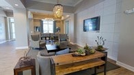 This sophisticated single-floor space seamlessly blends comfort and contemporary elegance in an open, flowing layout. The main lounge area is anchored by a rustic wooden coffee table and plush gray upholstered seating, creating an inviting gathering spot. A large wall-mounted flat-screen television is set against a striking tiled accent wall, adding a modern focal point to the room. Decorative greenery and sculptural arrangements lend a natural, curated touch to the space.

A graceful archway leads into a dedicated dining area, where a dark wood table is surrounded by coordinating chairs beneath a dazzling crystal chandelier that fills the space with warm, ambient light. Large windows dressed with soft teal drapes allow natural light to filter in, enhancing the bright and airy atmosphere throughout.

The space features crown molding, tray ceiling details, and recessed lighting that together elevate the overall sense of refinement. The neutral color palette of warm beiges and soft grays creates a cohesive, calming aesthetic that feels both polished and welcoming. Overall, this thoughtfully designed space offers a versatile and comfortable environment well-suited for relaxing, socializing, and dining.