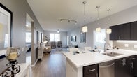 This thoughtfully designed single-floor residence showcases an open-concept layout that effortlessly connects the kitchen, dining, and living areas into one cohesive, flowing space. The kitchen features a generous white quartz island with bar seating, dark espresso cabinetry with sleek hardware, and elegant pendant lighting that adds a warm, contemporary touch. Track lighting overhead ensures the space is well-illuminated throughout the day. The dining area transitions naturally into a spacious living room, where large windows invite an abundance of natural light, creating a bright and airy ambiance. Neutral gray walls complement the warm wood-tone flooring that runs seamlessly throughout the entire space, tying each area together with a sense of continuity. The living room is equipped with a ceiling fan for added comfort and provides ample room for lounge seating and entertainment. The overall aesthetic is modern and clean, with a neutral color palette that offers a versatile backdrop for a variety of interior styles. The layout maximizes both functionality and openness, making this a welcoming and practical space suited for everyday living and entertaining alike.