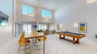Located at 3240 Champion Ring Rd in Fort Myers, Florida, this contemporary community clubhouse offers a well-appointed single-floor gathering space designed for both socializing and relaxation. The architecture is defined by dramatic double-height ceilings in the main area, allowing abundant natural light to pour in through large clerestory and floor-to-ceiling windows, creating a bright and airy atmosphere throughout.

The main lounge area features elegant marble-look tile flooring that flows seamlessly across the space, anchoring a seating arrangement of plush tan armchairs grouped around a stylish accent table — ideal for casual conversation or unwinding. Pendant drum lights hang from the soaring ceiling, complementing the clean white walls adorned with colorful contemporary artwork.

A long communal high-top table with bold yellow and orange bar chairs serves as a central hub for dining or collaborative gathering. Adjacent to it, a full-size pool table provides recreational entertainment. A kitchenette area, accented by a striking yellow backsplash wall and fitted with cabinetry and appliances, supports events and daily use.

A secondary lounge nook offers a more intimate setting with vibrant yellow accent chairs, a wall-mounted display, and direct access to an outdoor area. Additional corridors lead to private rooms with carpeted flooring, completing a versatile and thoughtfully designed community amenity space.
