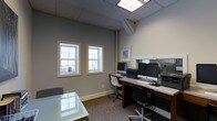 This single-floor professional office space offers a well-organized and functional environment designed to support focused work. The interior features a neutral color palette with soft gray walls and white trim, creating a calm and professional atmosphere throughout. A drop ceiling with recessed panel lighting ensures consistent, well-distributed illumination across the workspace. Two white-framed windows along the exterior wall invite natural light and provide views of the surrounding greenery, adding a refreshing contrast to the interior. The main workstation area is anchored by a long, dark wood desk that comfortably accommodates multiple computer workstations, including monitors, keyboards, and peripherals, making it well-suited for collaborative or multi-user tasks. Ergonomic rolling chairs complement the functional setup. A mirrored or glass-paneled surface along one wall adds depth and brightness to the room. A glass-topped table in the foreground provides additional workspace or meeting surface. Framed artwork on the wall contributes a touch of character to the otherwise clean and practical space. The carpeted flooring in a muted tone completes the professional aesthetic, offering comfort underfoot. Overall, the space balances practicality with a polished, welcoming office environment.