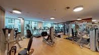 This fitness facility is thoughtfully designed to accommodate a full range of exercise needs within a single, open-plan floor. The space is divided into distinct functional zones, seamlessly connected by warm light hardwood flooring that runs throughout. A cardio zone along the perimeter benefits directly from floor-to-ceiling windows, offering outdoor views and flooding the area with natural light. This section houses multiple treadmills, elliptical trainers, stair climbers, and stationary bikes, all equipped with individual digital displays. Strategically placed mirrors expand the sense of space and assist users with form and technique.

The strength and resistance training zone is well-stocked with a variety of Hoist and other branded cable machines, weight benches, and functional training equipment, complemented by a dedicated free weights area with an organized rack of dumbbells and medicine balls. Rubber matting underfoot provides safety and stability in this zone.

A refined lounge area with a vertical wood slat partition wall adds a warm architectural detail, separating it from the workout floor. The lounge features a leather sofa, armchairs, and a solid wood coffee table beneath a statement pendant light, creating a comfortable space to rest and recover. Built-in shelving and a digital information display complete the facility's amenities.