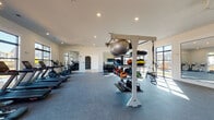 This modern fitness center is thoughtfully designed to accommodate a wide range of workout styles and fitness goals. The main exercise floor is covered in durable rubber flooring and is generously proportioned, offering ample room for movement between equipment stations. A row of treadmills and elliptical machines lines the perimeter, positioned to take full advantage of the large, black-framed windows that flood the space with natural light and provide views of the surrounding neighborhood. Ceiling fans and recessed lighting contribute to a bright, comfortable environment.

The functional training area is well-stocked with a multi-tiered equipment rack holding medicine balls, slam balls, stability balls, and kettlebells, alongside TRX suspension trainers. Full-length mirrors are strategically placed throughout to support proper form during workouts. A free weights section with dumbbells and weight benches adds further versatility to the strength training options.

Adjacent to the main floor is a dedicated studio room finished with light hardwood-style flooring and floor-to-ceiling windows that allow sunlight to pour in. This space is highlighted by a "Freedom Climber" rock climbing wall, offering a unique and engaging upper-body workout option. The studio's open layout makes it equally suitable for stretching, yoga, or group fitness activities.