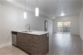 This well-appointed single-floor residence showcases a clean, modern aesthetic with light-toned luxury vinyl plank flooring flowing throughout the main living areas. The open-concept kitchen is a standout feature, centered around a large island with a white quartz countertop, an undermount sink, and wood-toned cabinetry. The kitchen is fully equipped with stainless steel appliances, including a refrigerator, electric range, and built-in microwave, complemented by white upper cabinets and a subway tile backsplash. Pendant lighting above the island adds a stylish focal point. The kitchen opens seamlessly into a bright, expansive living area filled with natural light from multiple windows fitted with blinds. Sliding glass doors lead to a private balcony with outdoor views, extending the sense of openness. The bathroom features a full soaking tub with a tiled surround, a curved shower rod, and a wood-toned vanity with a white countertop. The bedroom is generously sized and includes a walk-in closet outfitted with built-in shelving and dual hanging rods for ample storage. A dedicated in-unit laundry room with side-by-side washer and dryer and overhead shelving adds everyday convenience. Neutral tones and recessed lighting throughout create a bright, cohesive ambiance.