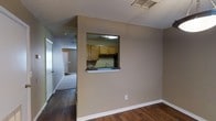 This two-floor space is designed with a practical and straightforward layout. The main living area features dark hardwood flooring that contrasts with the neutral beige-toned walls, creating a grounded and cohesive aesthetic. A textured popcorn ceiling adds a classic finish overhead, while white baseboards provide clean visual definition along the walls.

A standout architectural detail is the pass-through window cut into the wall between the living area and the kitchen, allowing for an open visual connection between the two spaces. The kitchen, visible through the pass-through, is equipped with medium-toned oak cabinetry, a sink, and dark countertops. An overhead light fixture illuminates the kitchen area, which also features a range hood above the cooking space.

A hallway extending from the living area leads further into the unit, where carpeted flooring transitions from the hardwood, suggesting a shift into additional rooms or private spaces. A door is visible at the end of the hallway, indicating access to additional areas of the two-floor layout. The overall ambiance is functional and understated, with a clean and move-in ready presentation.