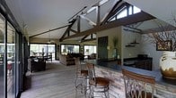 This single-story space makes a striking architectural statement with its soaring vaulted ceilings accented by bold, dark wood trusses that draw the eye upward and create a strong sense of height and grandeur. Track lighting runs along the ceiling lines, complementing two ceiling fans that serve both the bar and living areas. The open-concept layout connects the kitchen, bar, and living room in one expansive, cohesive space.

The bar area is a standout feature, finished with stacked stone veneer and topped with a sleek dark countertop. Cushioned bar stools with ornate metal frames line the bar, creating an inviting gathering spot. The kitchen flows naturally from this area, offering a functional yet stylish workspace.

The living area extends toward the rear of the space, furnished with comfortable seating and framed by large windows and sliding glass doors that allow abundant natural light to filter in. Views of an outdoor deck or patio are visible through the generous glazing, enhancing the connection between interior and exterior. Light wood-style flooring ties the entire space together, contributing to a warm, rustic-modern ambiance throughout.